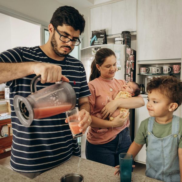Father pours juice while mother holds infant and toddler watches in kitchen