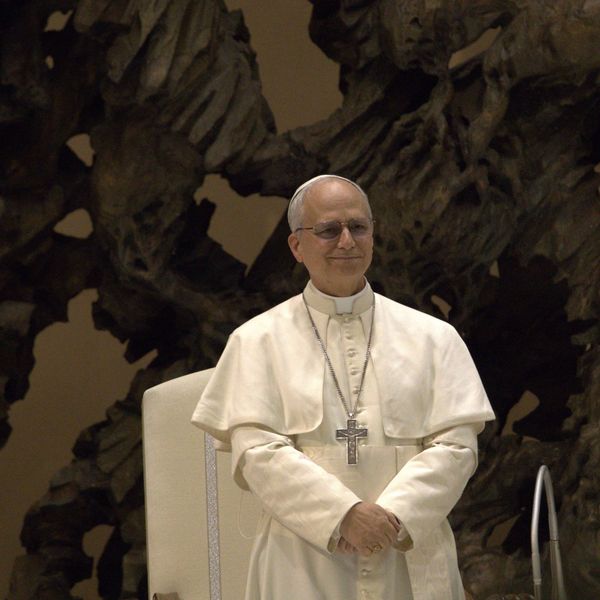 A pope in white robes stands smiling before a dark sculpture