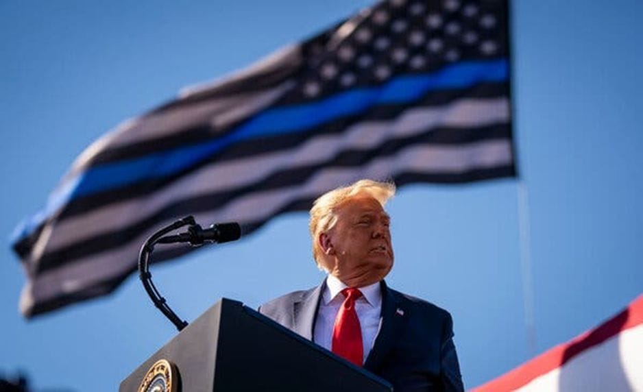 Donald Trump stands at a lectern in the foreground and looks off to the right. His urine-colored hand flaps atop his head. Fascist "blue line" American flag flaps in the background. Crisp blue sky.