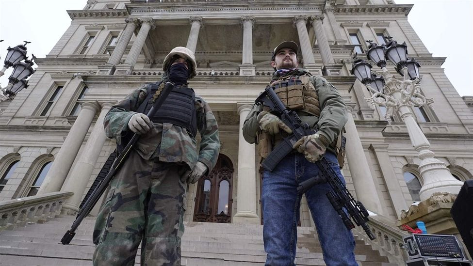 Two right wing extremist militia members, armed with massacre weapons and tactical gear, stand at the base of the stairs fronting the Michigan State Capitol, which they and their group have captured in order to demonstrate their exclusive right to not obey health ordinances during a pandemic.