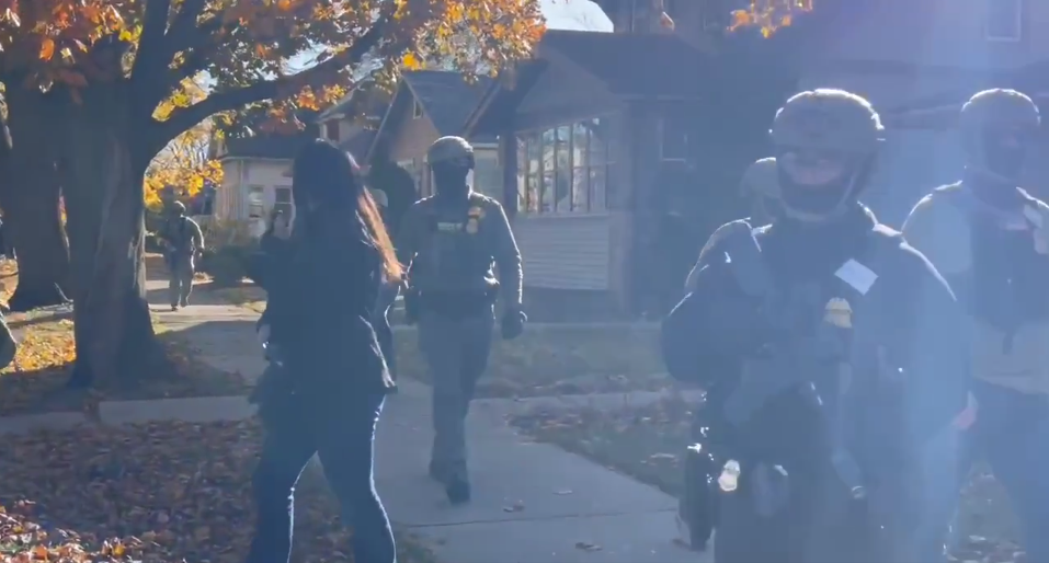 U.S. soldiers, or people posing as U.S. soldiers, in full tactical gear, wander a suburban U.S. street.