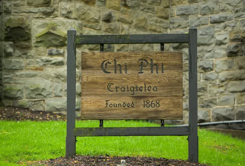A wooden sign in front of a stone wall. Gothic script reads "Chi Phi. Craigielea. Founded 1968."