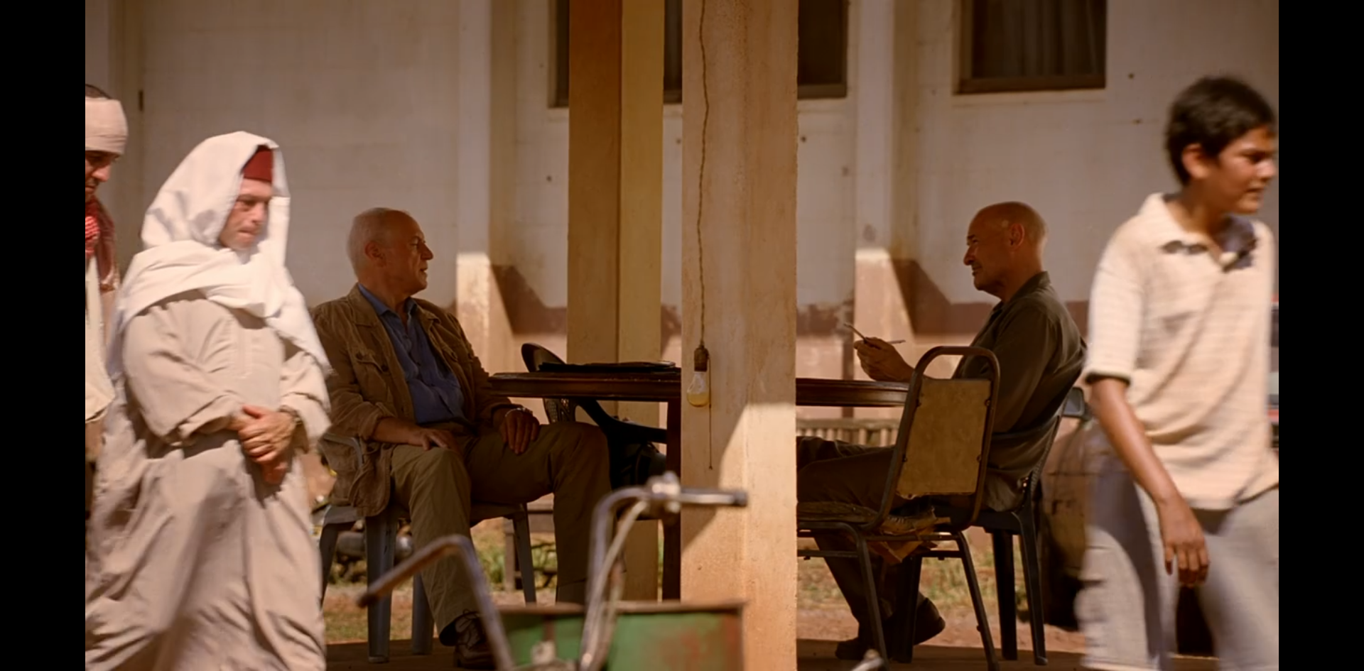 Widmore and Locke, seated, speak at an outdoor table.