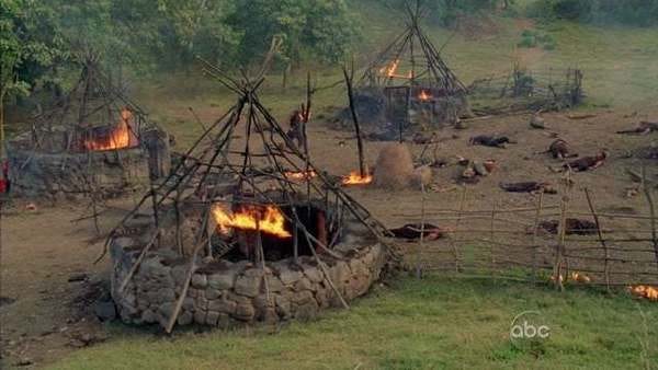 The remains of a village. Several huts are seen on fire, their roofs burned away. Several corpses are strewn around.