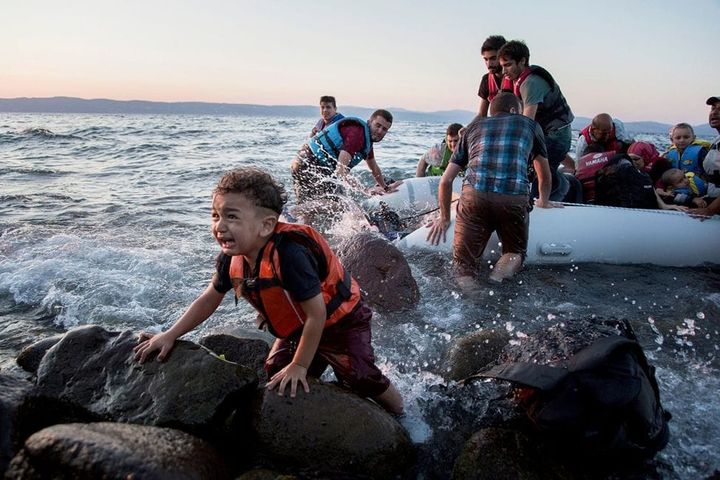 Refugees reach land in an inflatable raft. A young child in the foreground crouches on some rocks, crying.