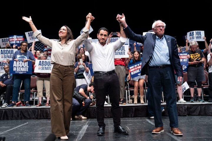 On a stage at a political rally, in front of a crowd of supporters, Abdul El-Sayed raises his hands, which are held by Alexandria Ocasio Cortez on his left and Bernie Sanders on his right.