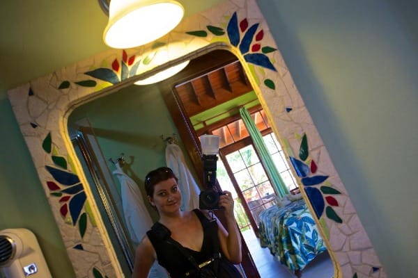 meg in a low cut black dress, holding a large DSLR camera with flash, in the mirror of a tropical-themed hotel room