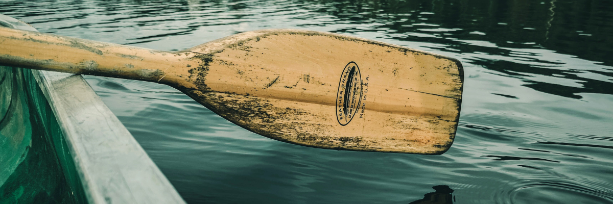 wooden oar dipping into calm water from inside of a boat