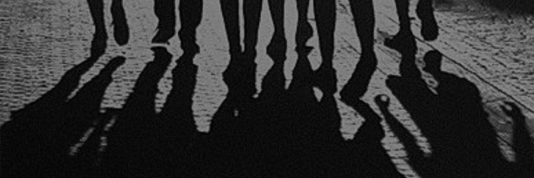 black & white photograph of a group of people walking, only their feet visible as well as the long shadows they're creating togethe