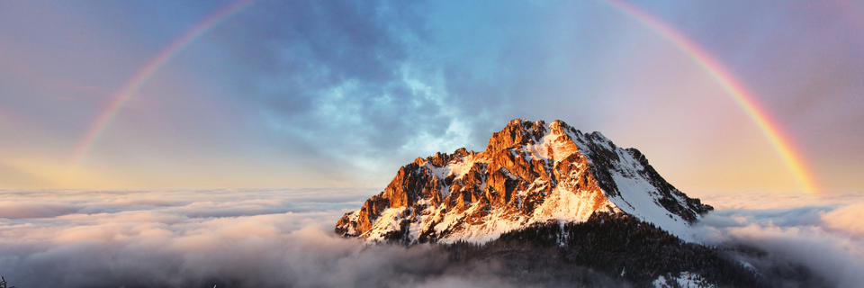 rocky peak of a mountain bursting through clouds, with sky and a rainbow stretching overhead