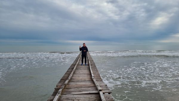 A wooden jetty stretching out into the sea under a heavy grey sky, with a lone figure standing near the far end.