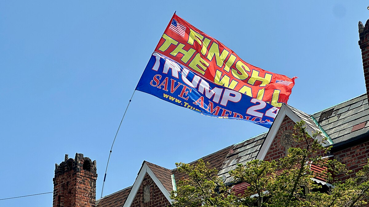 A flag reading "Finish the Wall trump 24 Save America" waving in front of a house