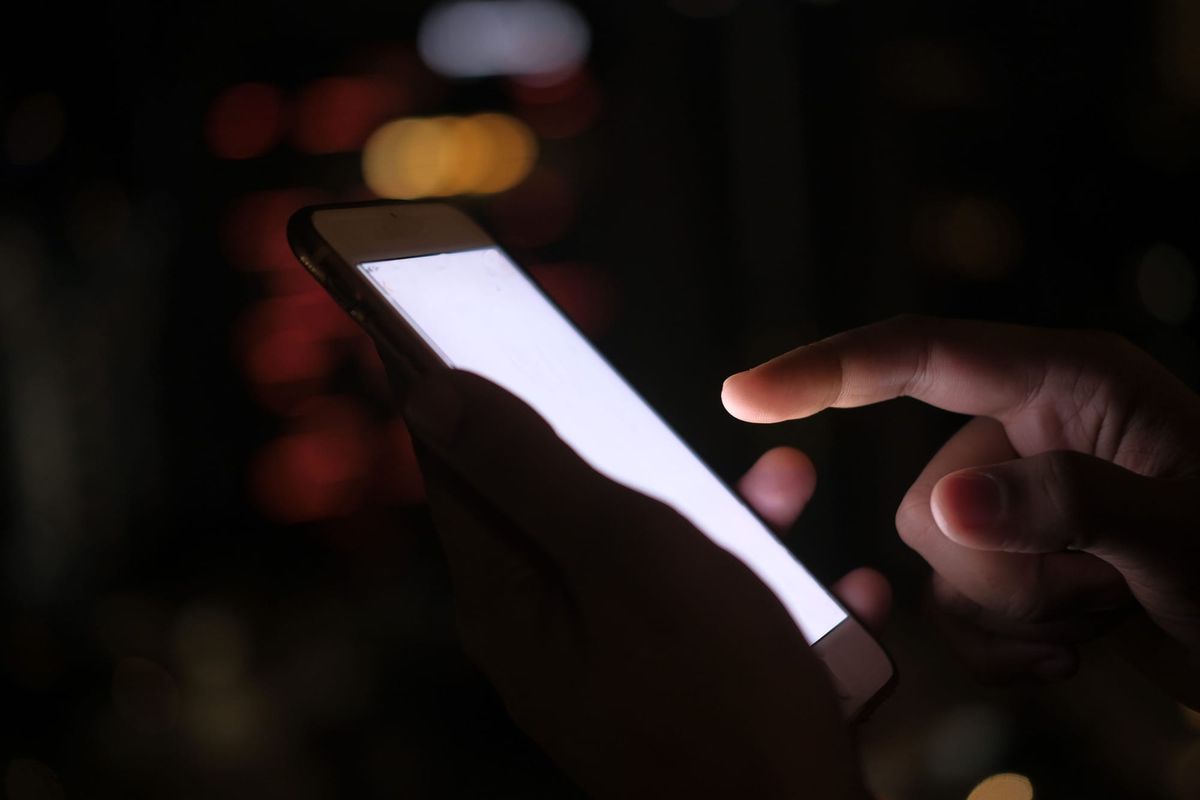 Close-up of a hand using a smartphone in the dark with blurred city lights in the background.