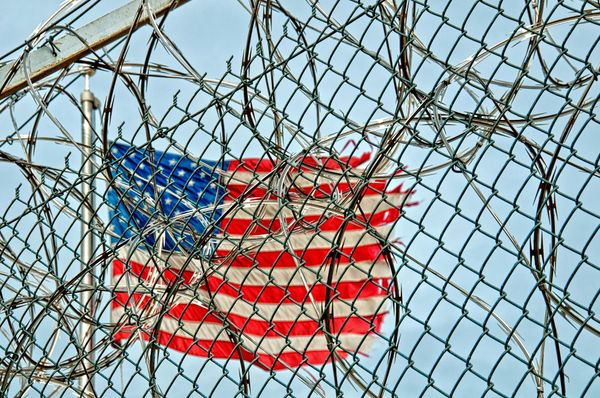 A tattered USA flag flies on a pole behind barbed wire and a chainlink fence.