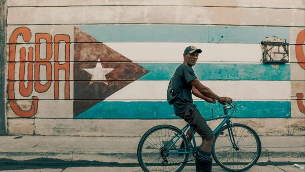 A man rides a bicycle by a vibrant Cuban flag mural in Havana.