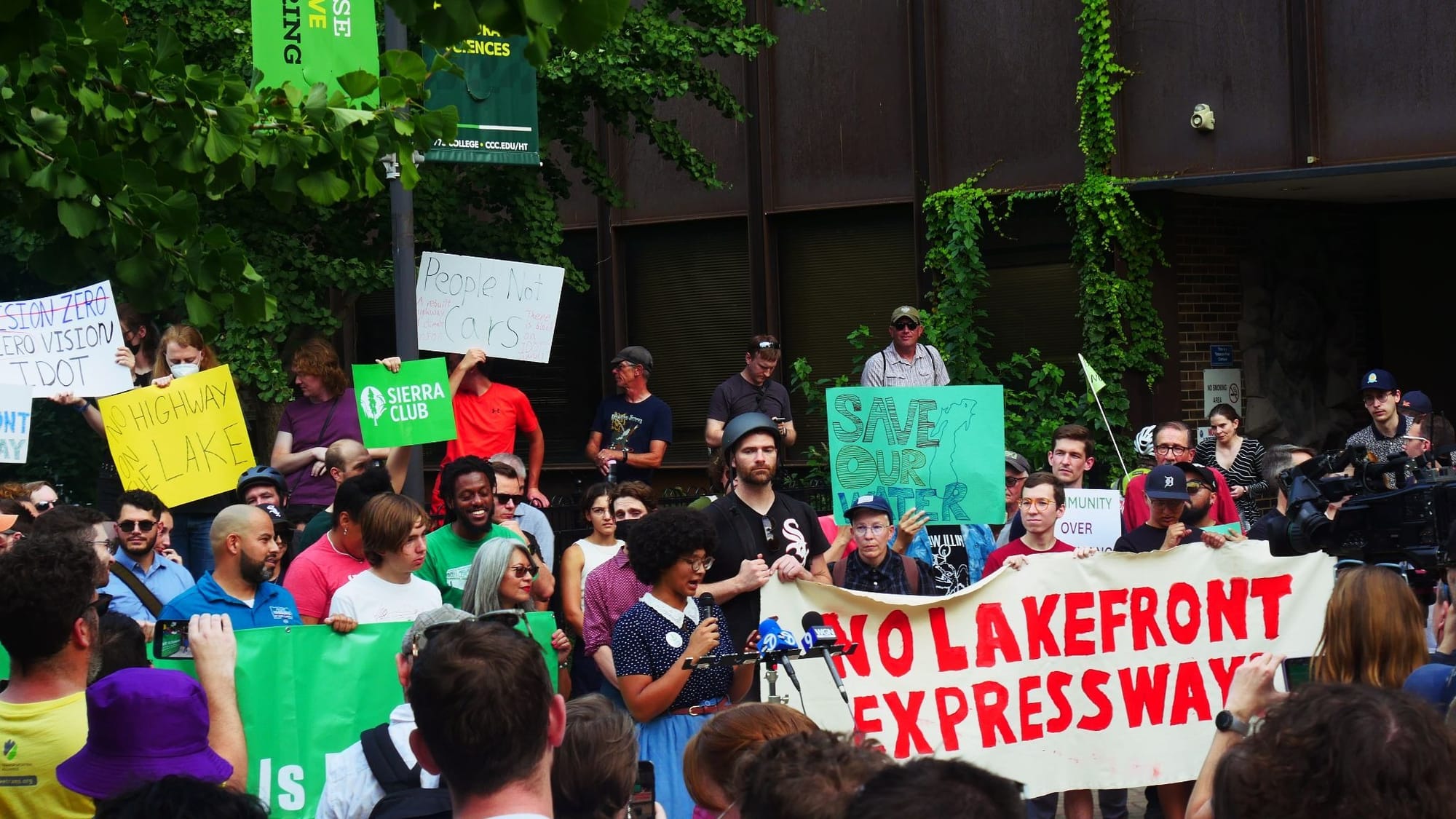 At a protest rally, a person speaks into a row of news microphones in front of a banner reading "No Lakefront Expressway".