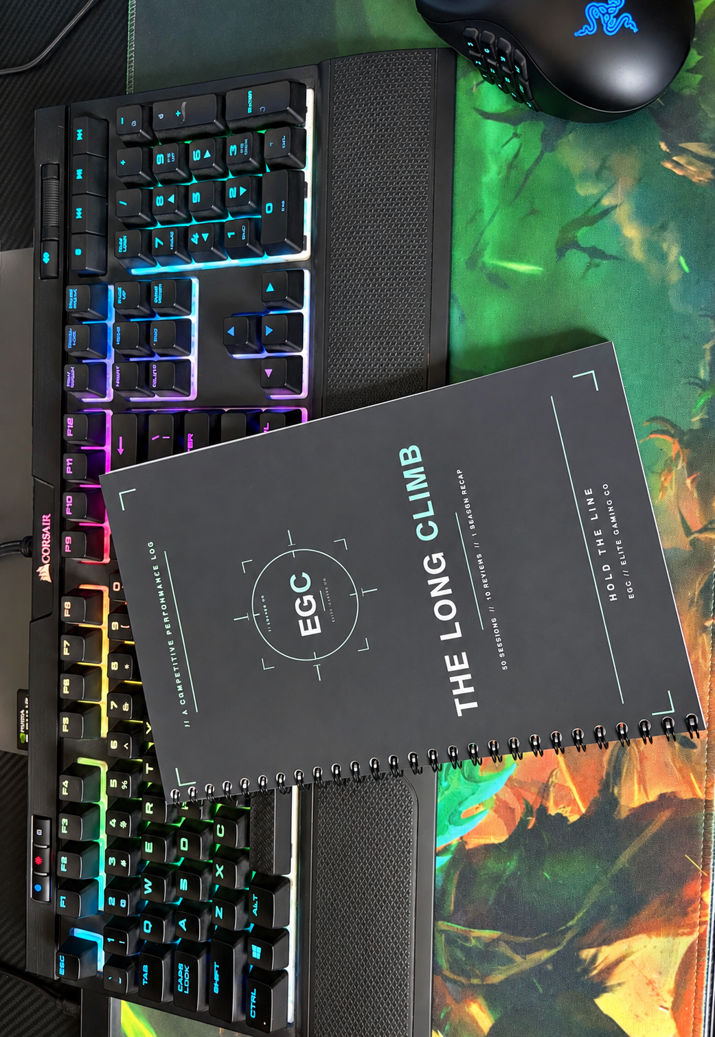 The Long Climb spiral-bound performance log on a gaming desk beside an RGB keyboard.