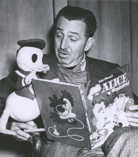 A black-and-white photo of Walt Disney reading Alice in Wonderland to a Donald Duck puppet