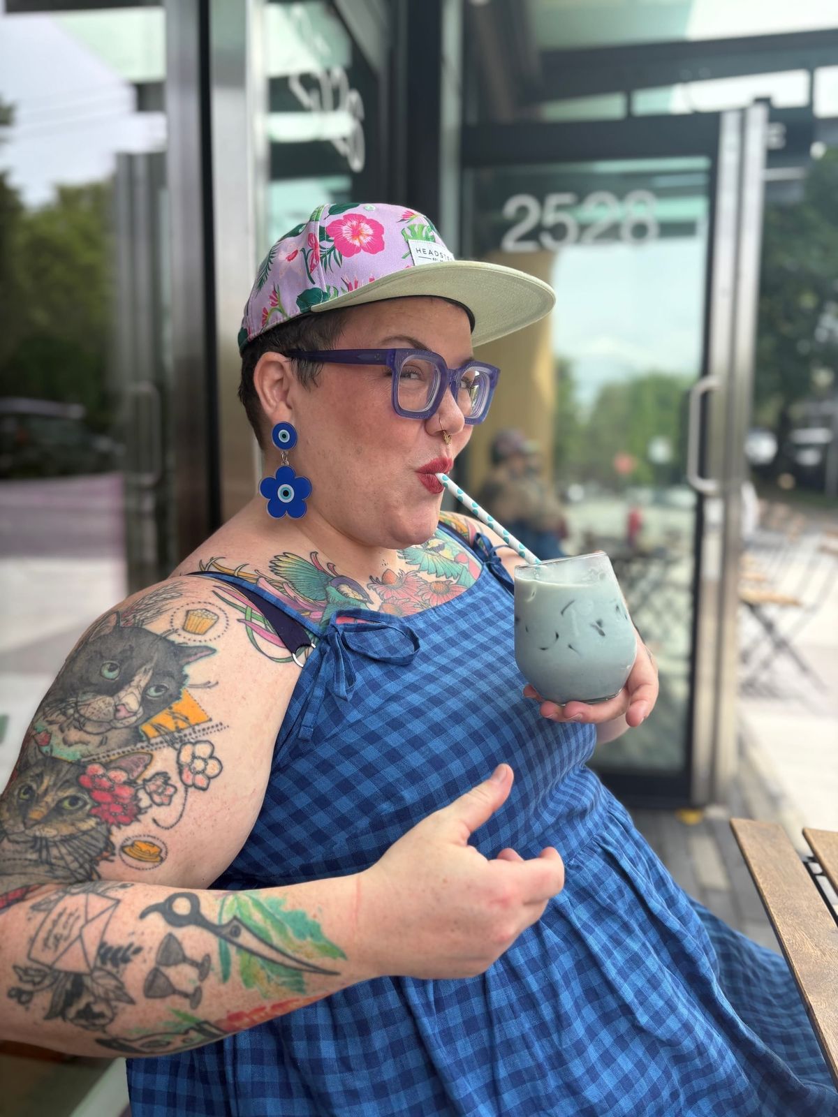 Photo of Hannah sitting on a patio drinking a blue iced latte. They have chunky purple glasses, blue Nazar earrings, a blue gingham dress, a floral baseball cap, and many colourful tattoos.