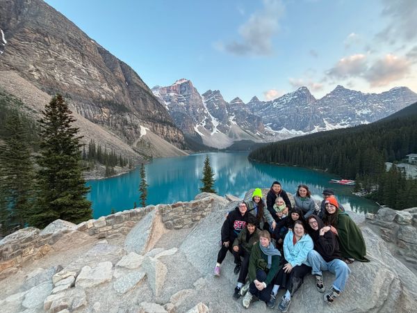 12 people, including Hannah, sit on a rocky outcropping overlooking a bright blue lake surrounded by mountains