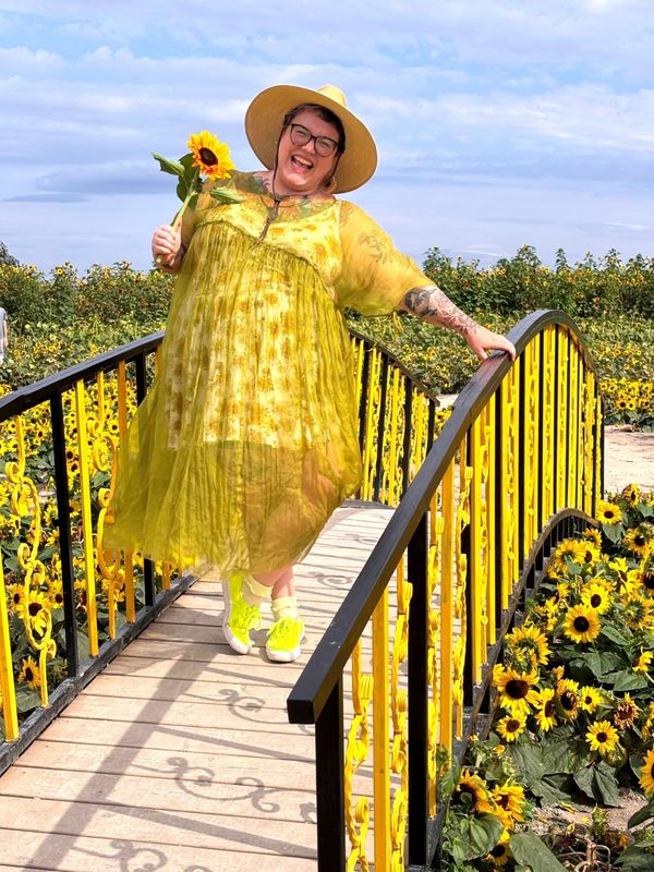 Hannah is standing on a yellow and black bridge in a field of sunflowers, wearing a sunflower dress, holding a sunflower, grinning.