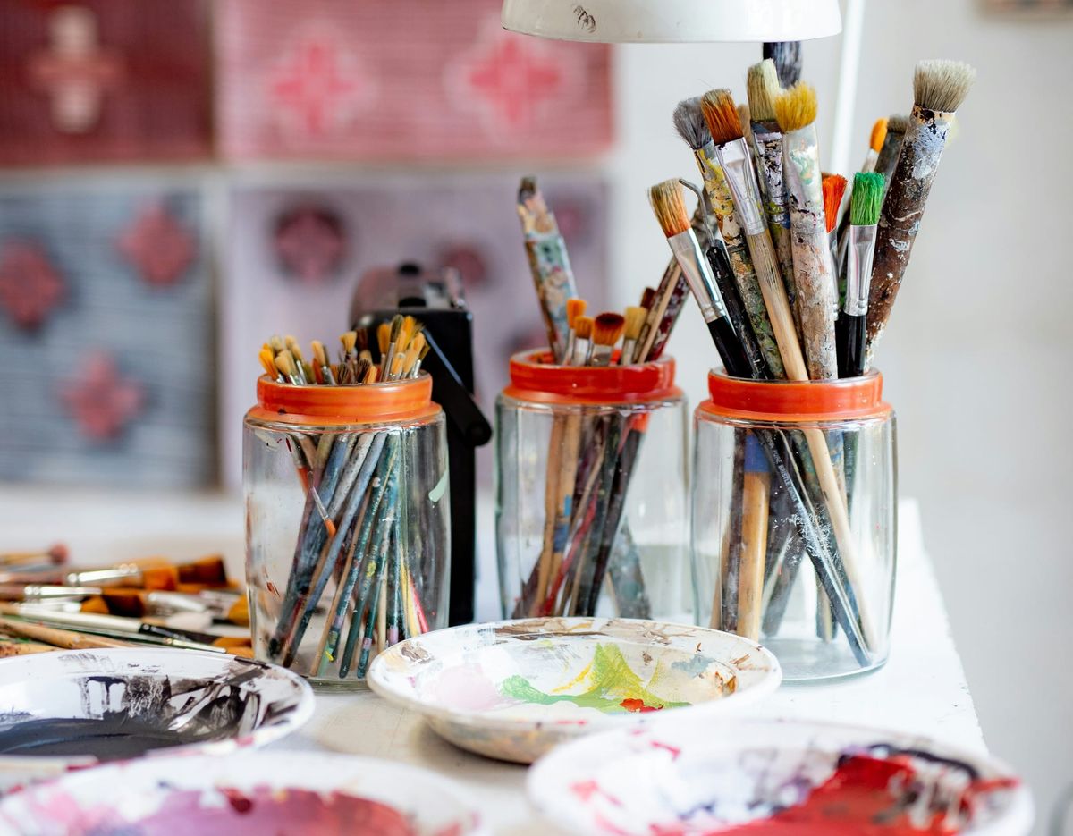 Art supplies in foreground and finished pieces in same color palette hanging on wall in background