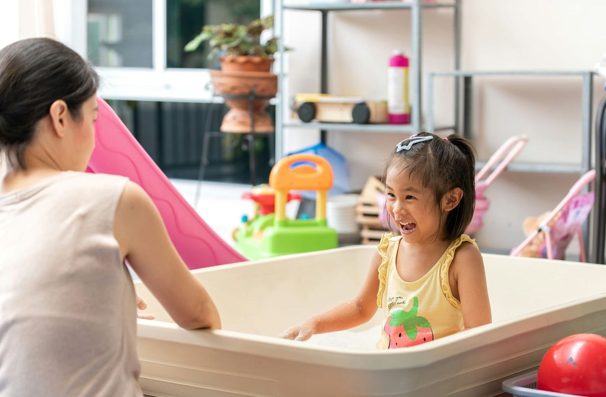A girl with dark hair and a yellow tank top plays in a large white indoor sandbox with an adult nearby.