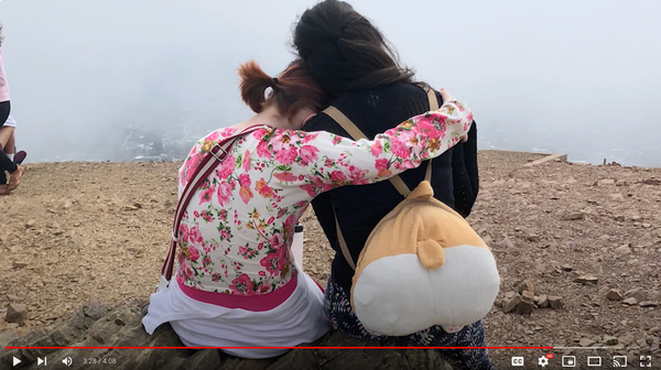 A teenager and a young adult sit in an embrace facing away from the camera, at the summit of a hill with fog in the distance