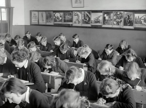 Black and white photo of white children in school uniforms bent over written coursework