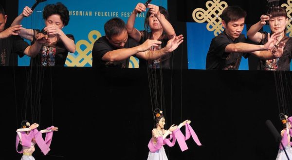 Six performers control three marionettes clothed in pink and purple dresses and ribbons