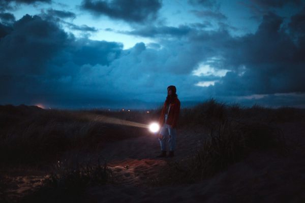 A person walking at dusk, perhaps on or near a beach, shining a flashlight