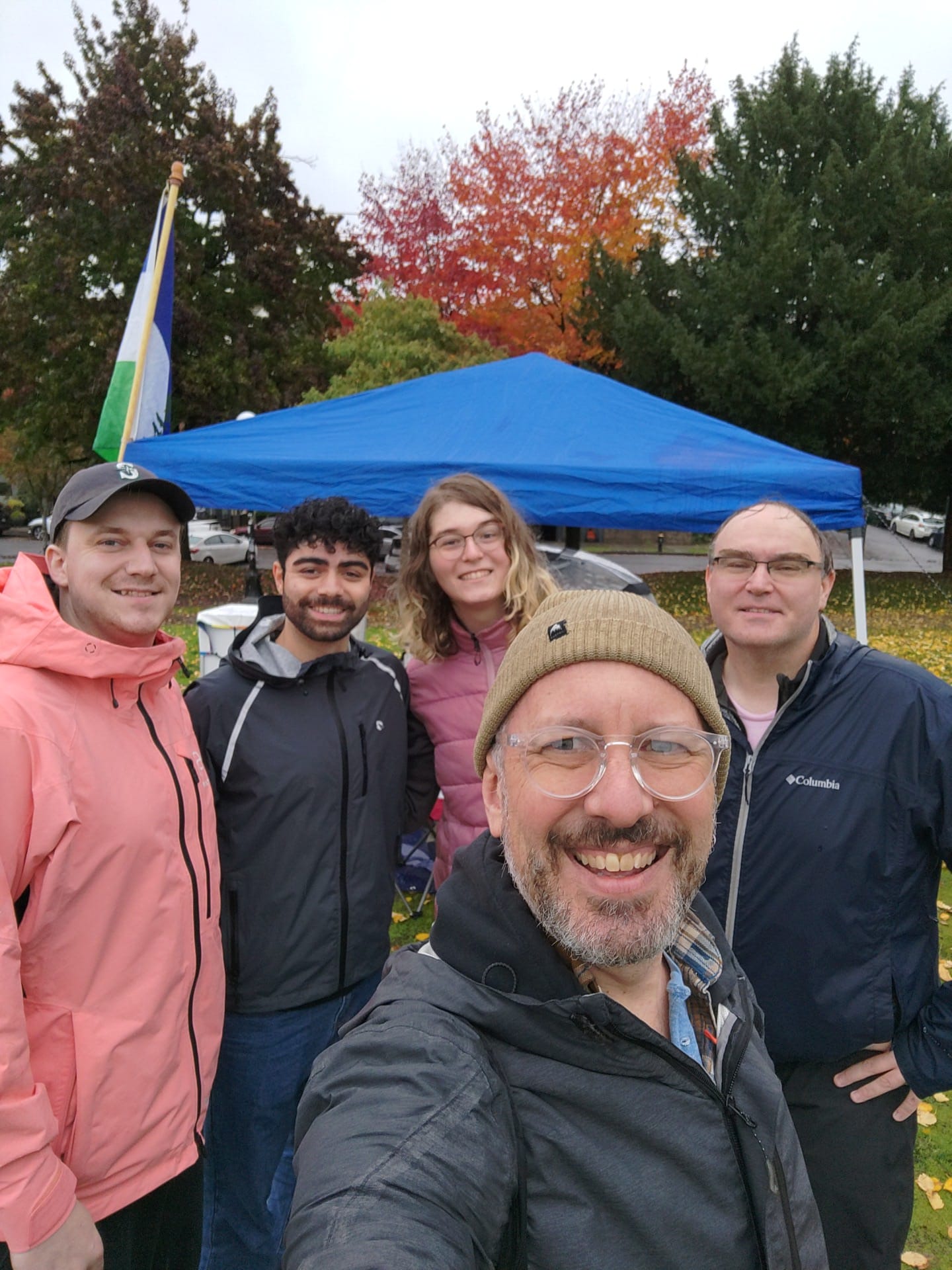 Several people stand smiling in front of a rain canopy and a Cascadia flag.