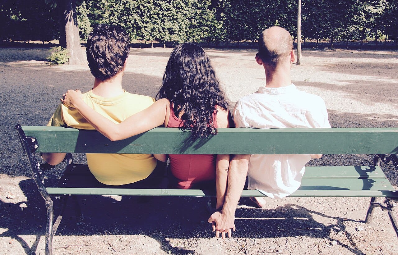 Three people sit on a bench, one with her arm around one person and holding hands with another