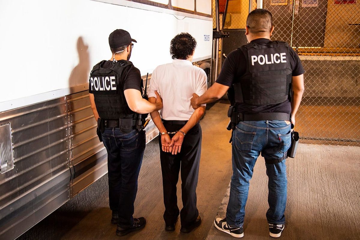 A man is taken into custody by two ICE agents with POLICE on the back of their uniforms.