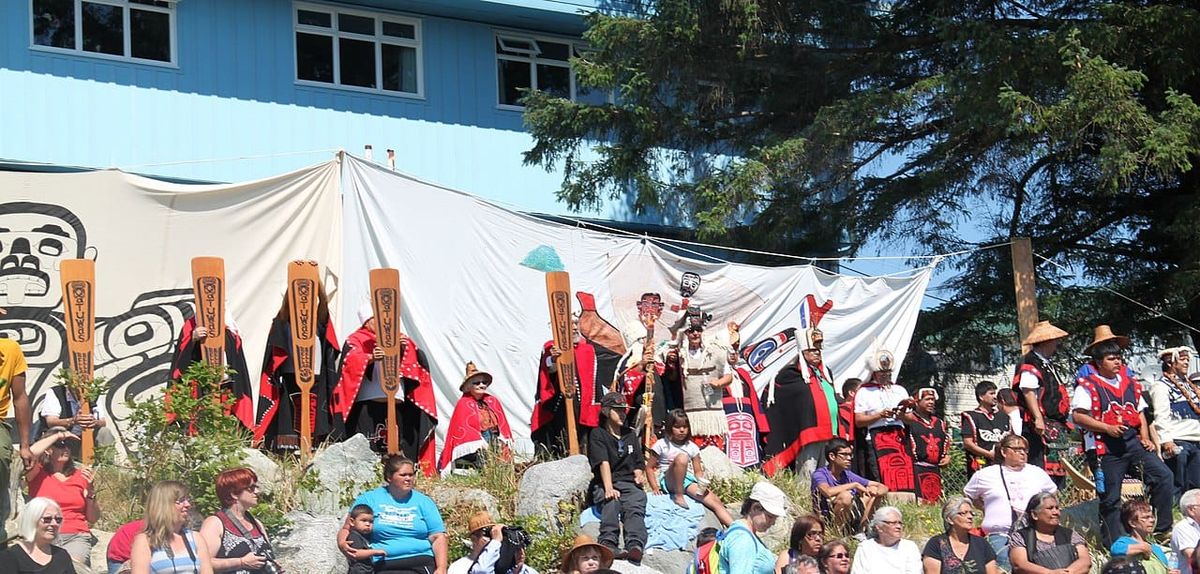 Members of the Heiltsuk First Nation gather wearing traditional red and black shell blankets, cedar hats, and holding traditional canoe paddles.
