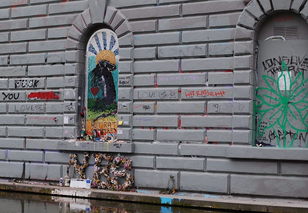 A mosaic of a rat, with rising sun and adorned with trans flags, candles, and a sculpture that reads YES.