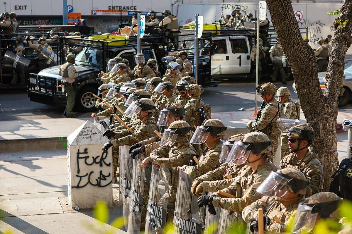 National guard troops in combat fatigues with plastic shields and batons stand in a line next to a garbage can spray painted with the words Fuck ICE.