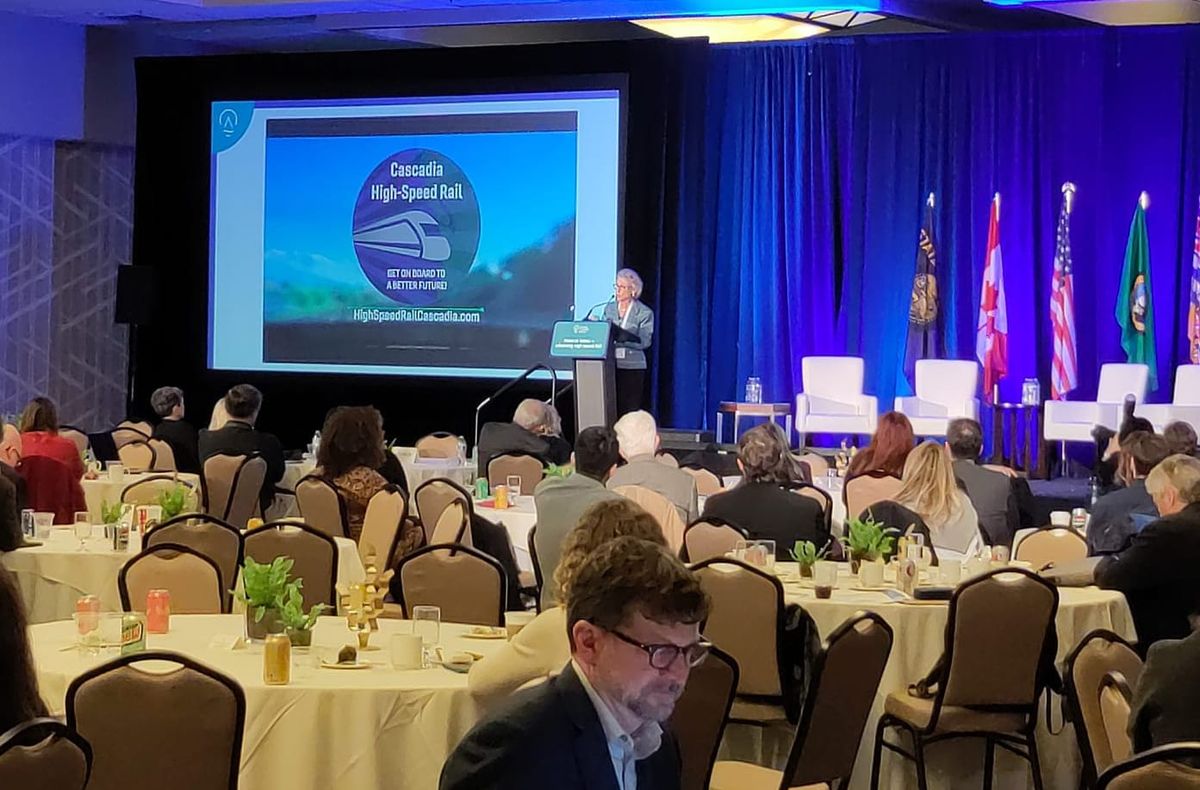 A speaker addresses a ballroom filled with various regional leaders. A power point reads Cascadia High Speed Rail. 