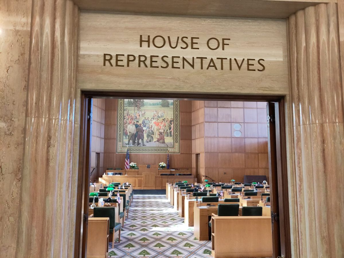 Desks and main podium of the Oregon state house seen through a doorway with a sign reading House of Representatives above the door.