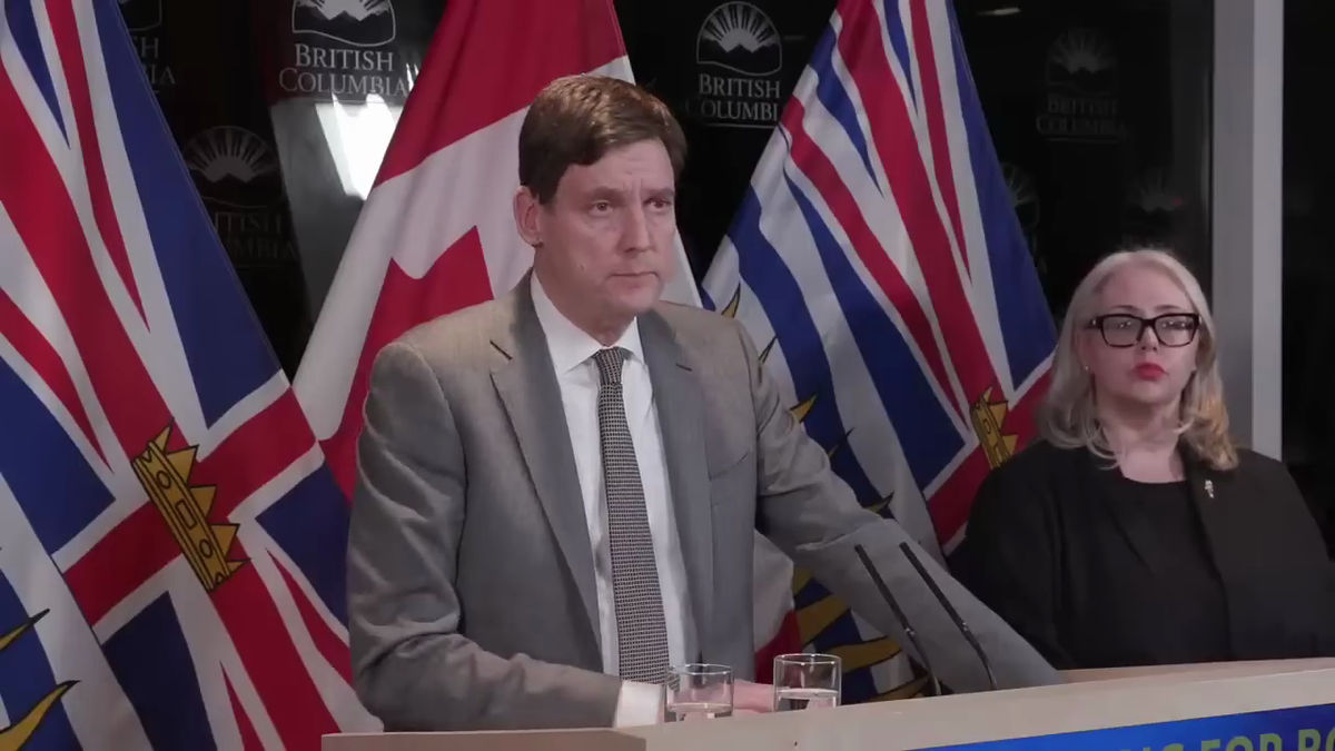 A man in a suit and a woman in business attire speak to the public in front of Canadian and BC flags.