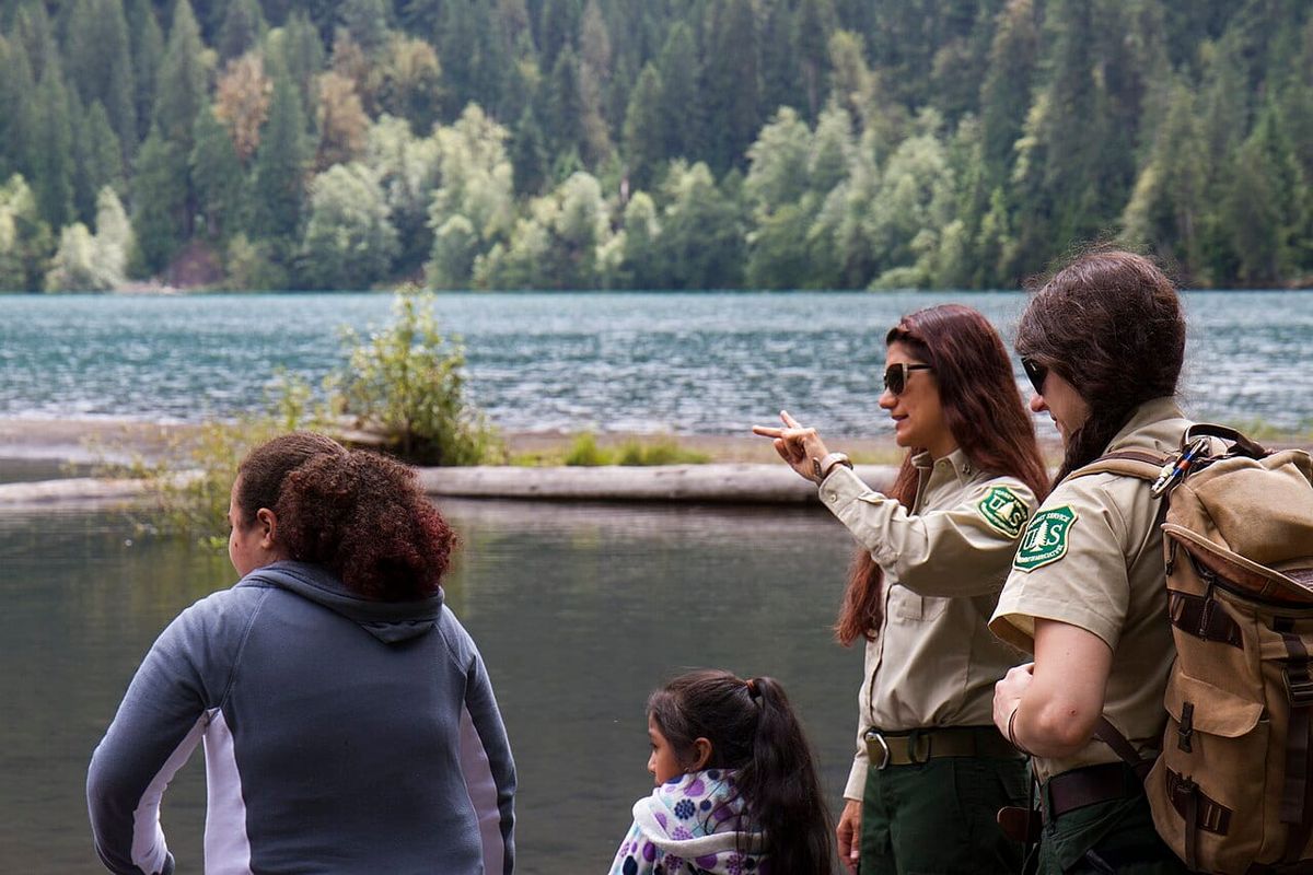 Two female forest rangers talk about nature to two girls on the shore of a mountain lake surrounded by evergreen trees