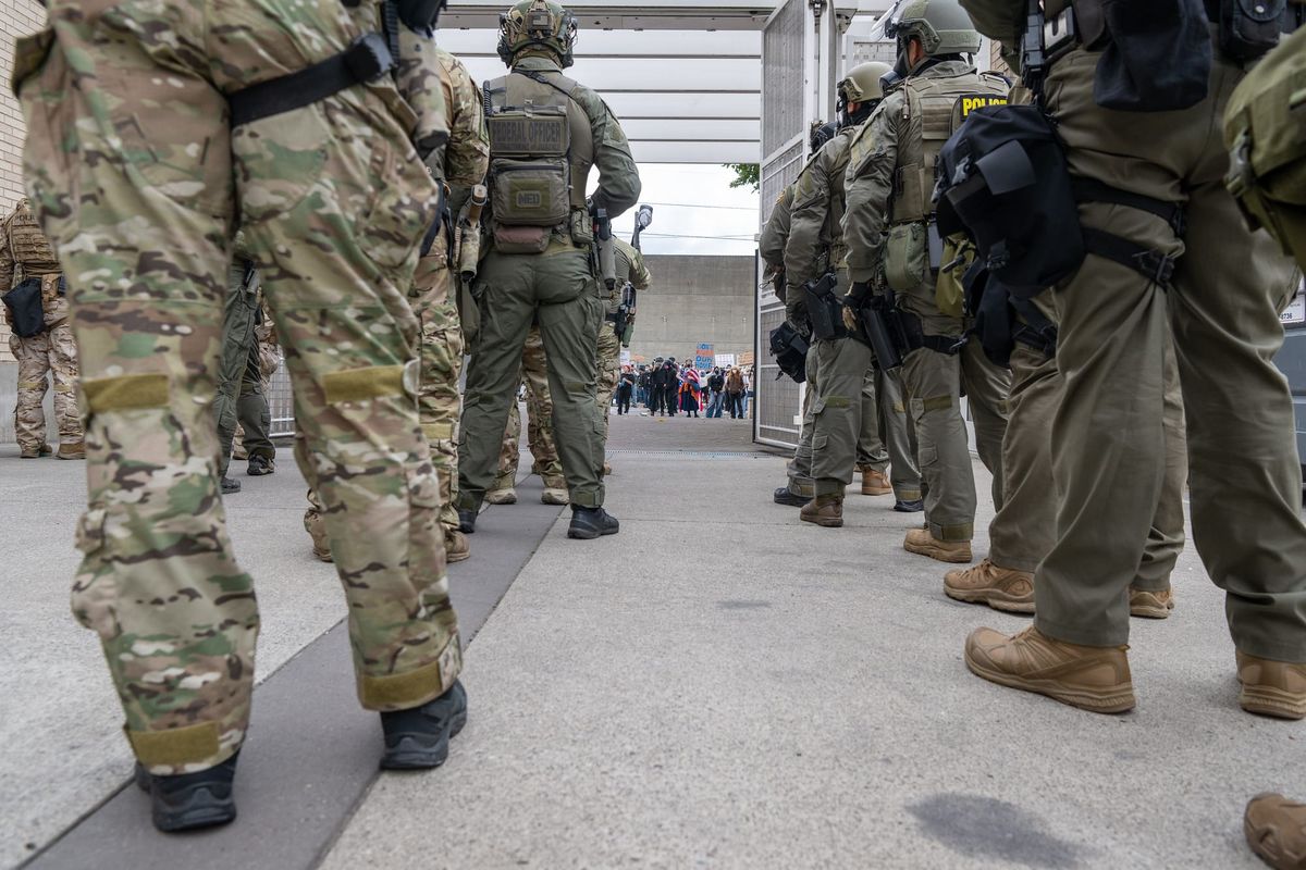 Well armed goons in camoflauge and tactical gear face a small crowd of protesters.