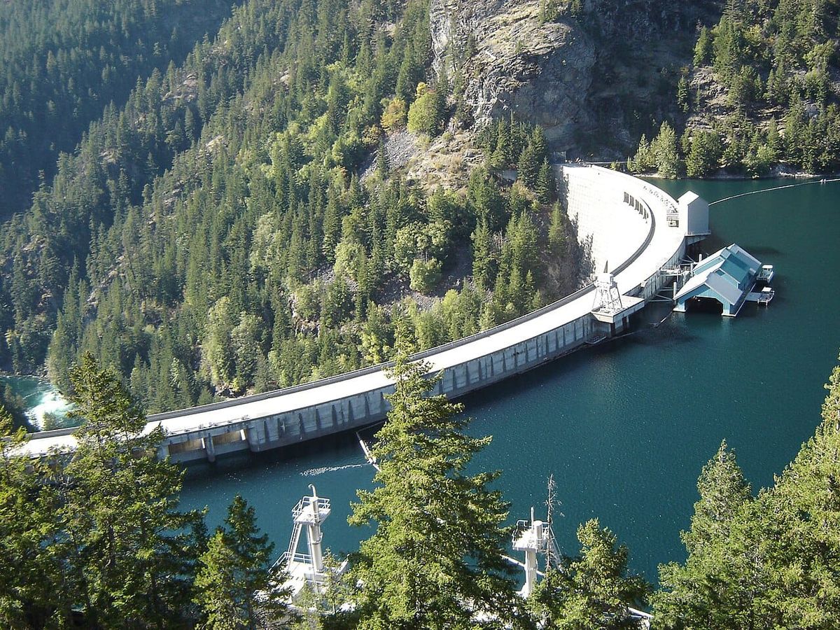 A deep blue lake behind a wide curving dam surrounded by steep mountainsides covered in evergreen trees