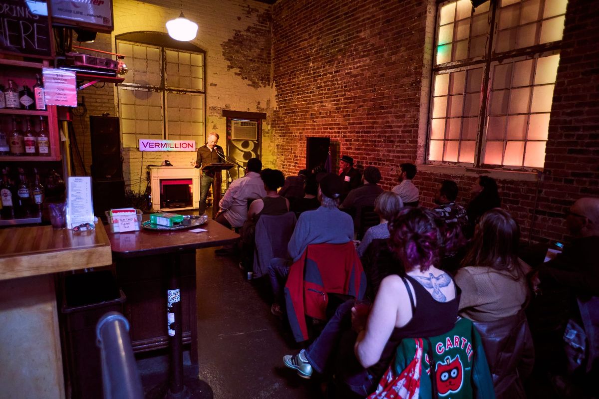 A guy stands behind a podium in a brick-walled bar in front of a full crowd of Cascadia supporters.