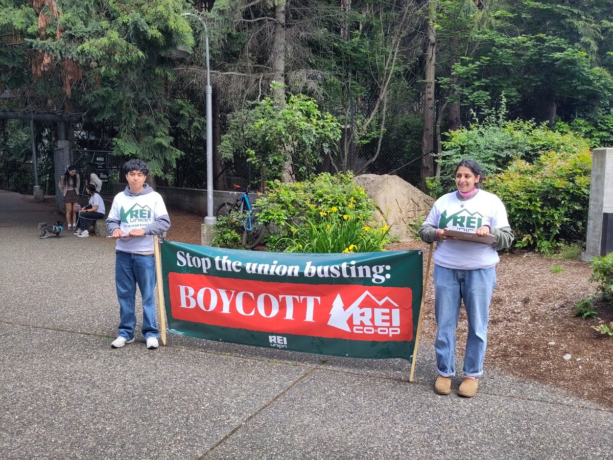Two organizers wearing REI Union shirts hold a sign that says: Stop the union busting: boycott REI co-op in front of a forested store entrance