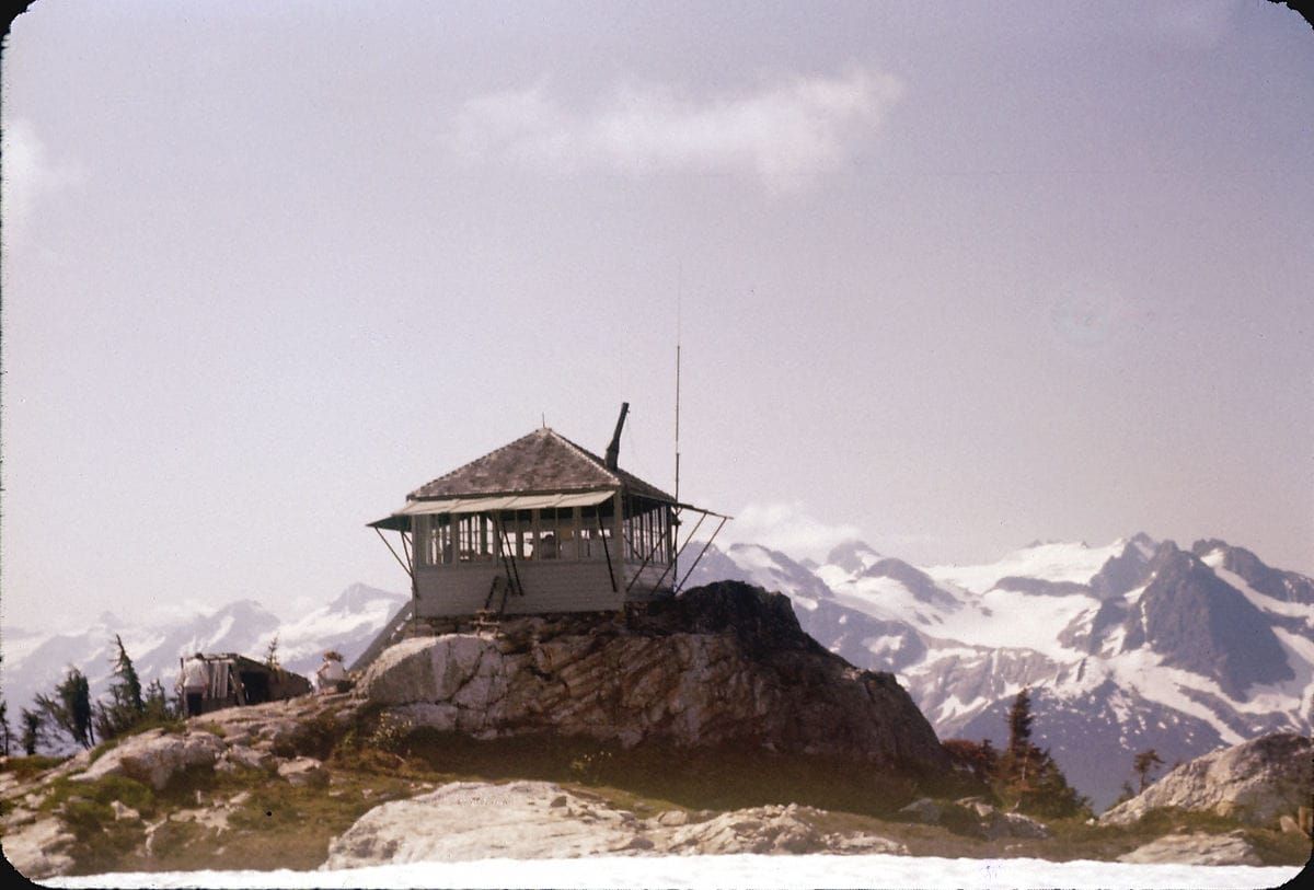 A forest lookout cabin on top of a mountain, with glaciated peaks in the distance.