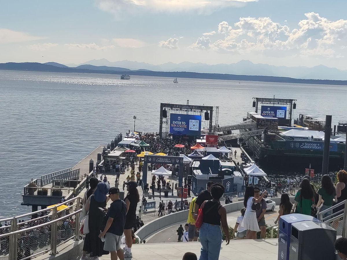 A view of a pedestrian walkway, a public TV screening area on a pier, and a vast bay with mountains and a ferry boat in the distance
