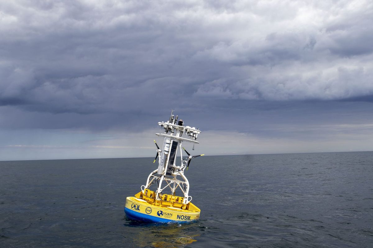 A scientific buoy with an array of monitors on top of it is seen in a wide ocean under a stormy sky.
