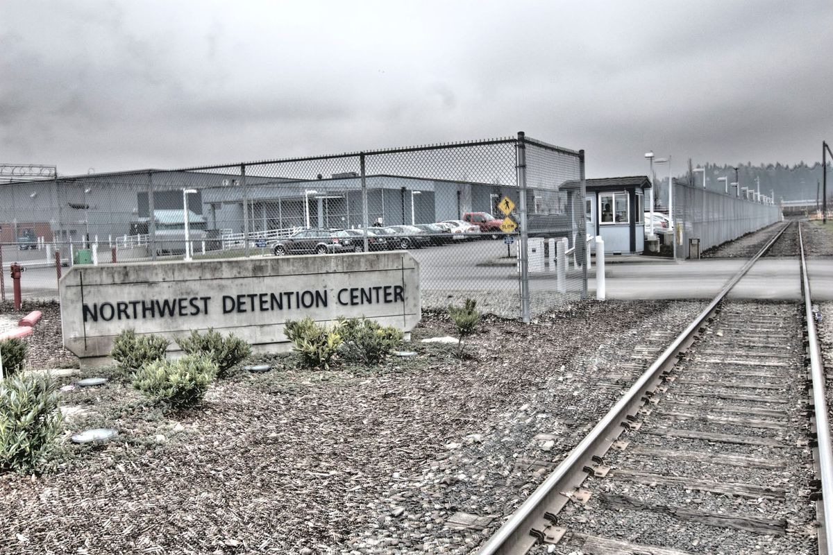 A jail behind barbed wire near train tracks and a sign that reads Northwest Detention Center.