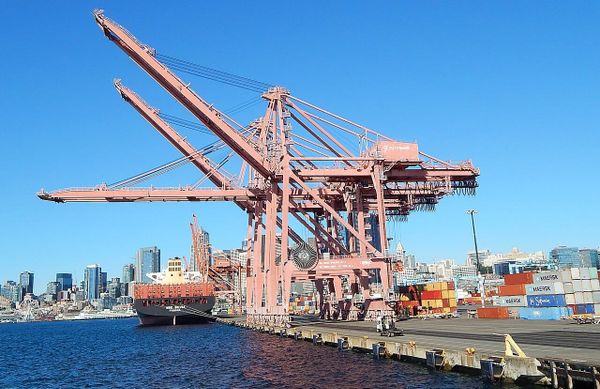 Large cranes load and unload containers above a cargo ship, with the Seattle skyine in the background.